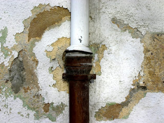 A white pipe with rusty paint on the wall, indicating water damage to the plumbing system.
