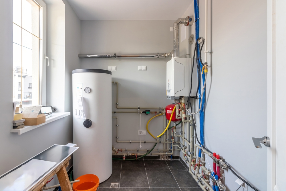 A utility room featuring a white water heater alongside various pipes and a gas boiler. A window allows natural light to illuminate the tiled floor, where a small orange bucket rests. The walls are painted in a soothing light gray tone.