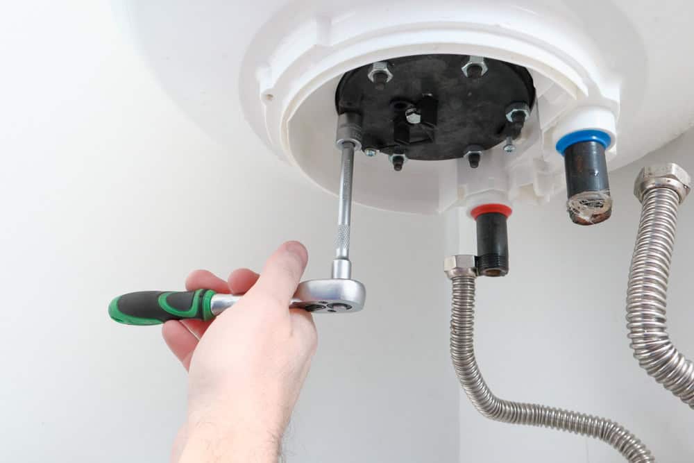 A hand uses a wrench to adjust a nut under a water heater. Metal pipes with various fittings are visible, and the wrench has a green and black handle. The focus is on the underside of the water heater.