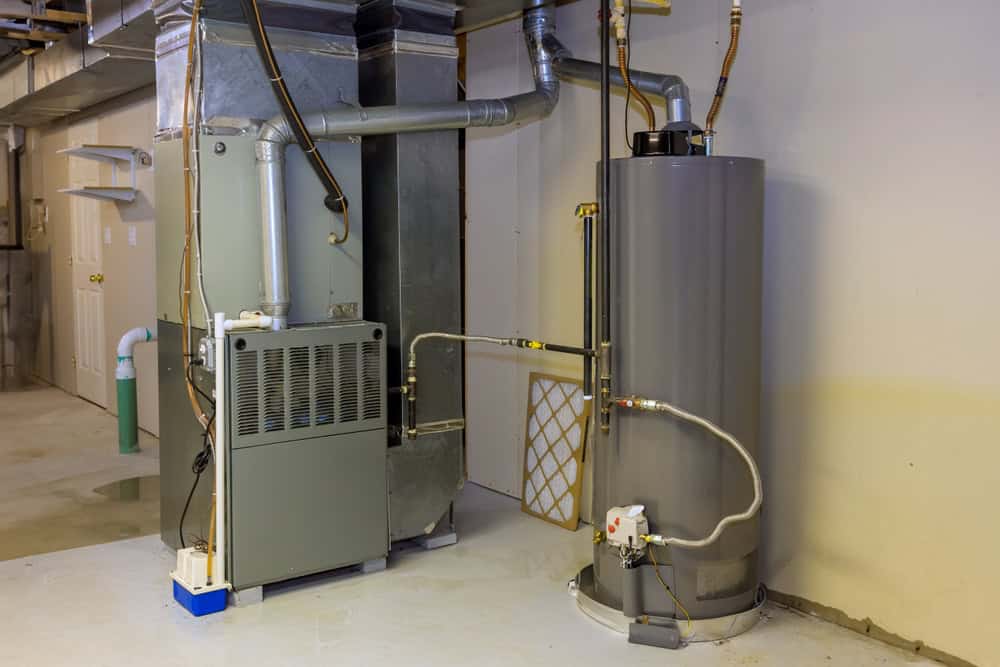 A basement with a gray furnace on the left and a tall, cylindrical water heater on the right. The furnace has vents and pipes connected, while the water heater is linked by pipes and has a valve. Walls are light-colored and the floor is concrete.