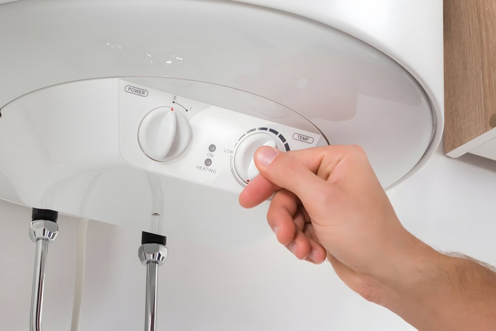 A hand turning the temperature control knob on a white water heater. The panel has a power button, a temperature dial, and labels indicating "Low" and "Heating." Two metal pipes are visible below the heater.